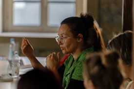 Eine Frau mit braunen Haaren, Brille und einem grünen Hemd sitzt am Tisch und gestikuliert erklärend