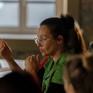 Eine Frau mit braunen Haaren, Brille und einem grünen Hemd sitzt am Tisch und gestikuliert erklärend