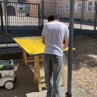Person mit weißem T-Shirt und Jeans steht an einem gelben Tisch unter einem Sonnenschutz im Außenbereich