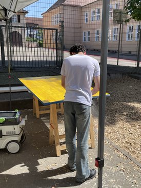 Person mit weißem T-Shirt und Jeans steht an einem gelben Tisch unter einem Sonnenschutz im Außenbereich