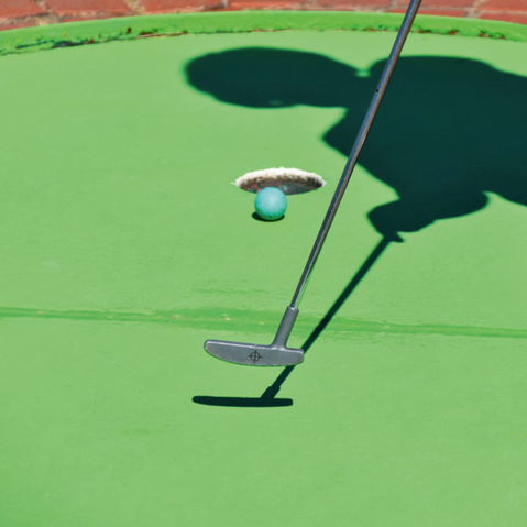 Minigolf-Putter trifft blauen Ball auf grünem Spielfeld mit Schatten einer Person im Hintergrund, dargestellt in einem kreisförmigen Ausschnitt, der wie ein Loch wirkt.