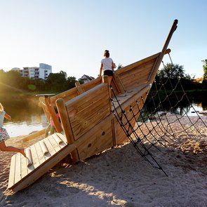 Blick auf ein Spielgerüst in Bootsform am Neckarstrand. Zwei Kinder spielen darauf.