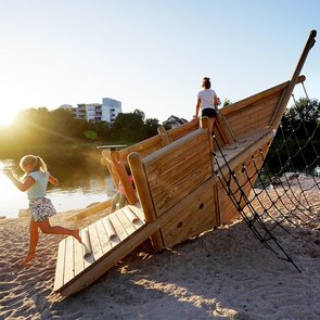 Blick auf ein Spielgerüst in Bootsform am Neckarstrand. Zwei Kinder spielen darauf.