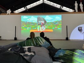 Zwei Personen sitzen auf großen Kissen und schauen ein Videospiel auf einer Leinwand beim Kunst im Game Symposium an.