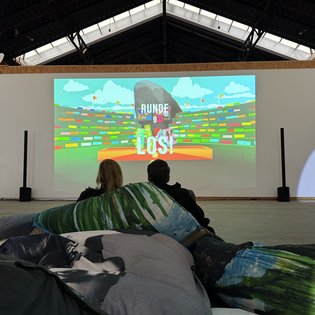 Zwei Personen sitzen auf großen Kissen und schauen ein Videospiel auf einer Leinwand beim Kunst im Game Symposium an.