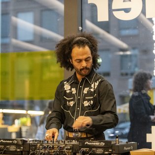 DJ Enkidu am Mischpult. Er hat längeres, lockiges Haar, das mit einem Band zurückgebunden ist und trägt ein schwarzes Hemd mit silbernen Details.