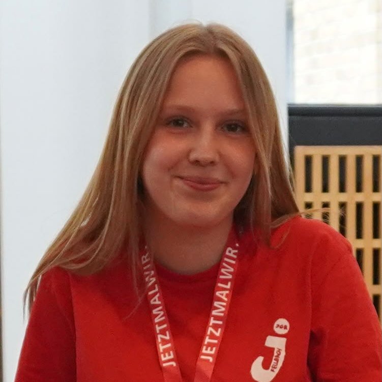 Junge Frau mit langen blonden Haaren trägt rotes T-Shirt und rotes Lanyard mit weißer Aufschrift