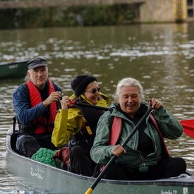 Ein mit Elisabeth Maier und zwei weiteren Personen besetztes Kanu, das auf einem Gewässer fährt. Alle drei Personen tragen Schwimmwesten und halten ein Paddel in den Händen.