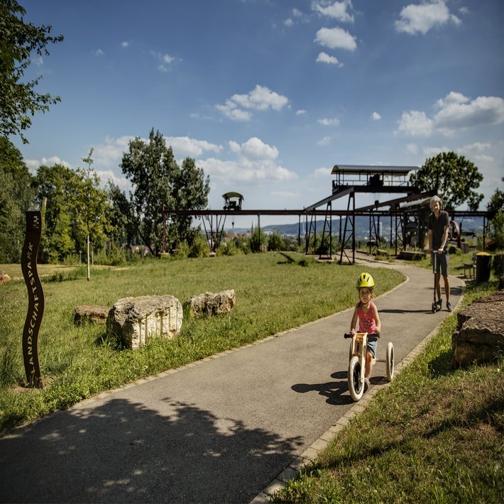 Bild: ein kleines Kind fährt mit einem Dreirad auf einem Weg durch einen Park. Ein Mann, vermutlich der Vater, fährt mit dem Roller hinter dem Kind. Im Hintergrund sind alte Bergbauanlagen zu sehen und es eröffnet sich ein Blick in ein Tal.