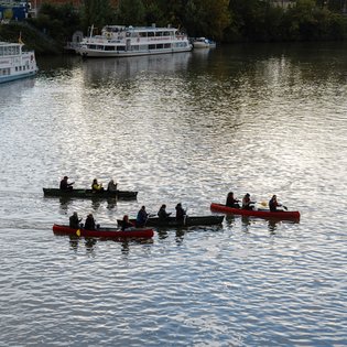 Vier mit Personen besetzte Kanus, die nah aneinander auf einem Gewässer fahren. Im Hintergrund sind größere Schiffe zu sehen.