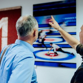 Zwei Männer betrachten ein Bild an der Wand, das eine Curling-Szene zeigt, einer der Männer zeigt mit dem Finger darauf
