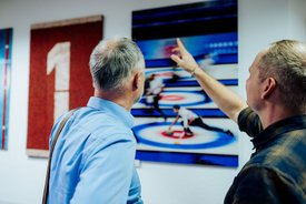 Zwei Männer betrachten ein Bild an der Wand, das eine Curling-Szene zeigt, einer der Männer zeigt mit dem Finger darauf