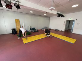 Zwei Personen knien auf rotem Boden und arbeiten an einem großen gelben Minigolfhindernis aus Holz in einem hellen Raum mit weißen Wänden und Deckenleuchten.