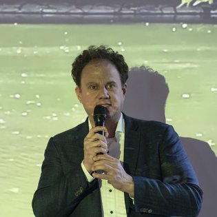 Dr. Matthias Knecht, OB Ludwigsburg und Vorsitzender der KulturRegion Stuttgart, während seiner Eröffnungsrede. Er hält ein Mikrofon in beiden Händen. Im Hintergrund auf einer Leinwand: ein Bild eines Gewässers.