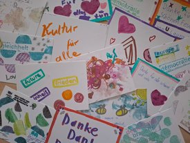bemalte Postkarten auf denen bunte Zeichnungen und Stempelpoesie stehen: "Demokratie braucht Utopie" oder "Kultur für alle" oder auch "Danke" oder "Be bautiful"