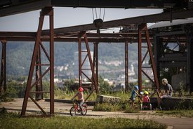 Kinder fahren Fahrrad zwischen stillgelegten Bergbauanlagen im Travertinpark. Ein Mann, vermutlich der Vater, sitzt auf einer Bank und schaut den Kindern zu. Im Hintergrund öffnet sich der Blick auf ein Tal.