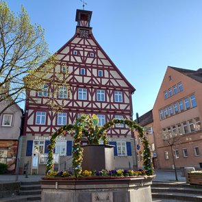 Fachwerkhaus mit Uhrturm hinter einem Brunnen, der mit Blumen und grünen Bögen geschmückt ist, an einem sonnigen Tag auf einem Marktplatz