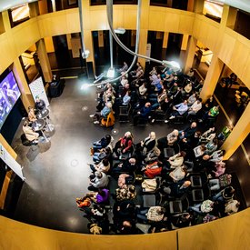 Bild der Podcast-Aufnahme im Foyer des StadtPalais, fotografiert von der Galerie im ersten Stock des Gebäudes. Links im Bild sitzen der Moderator und die beiden Gäst*innen auf Barhockern an Tischen vor einer Videoleinwand auf der Bilder gezeigt werden. Rechts das Publikum in Stuhlreihen mit einem Mittelgang.