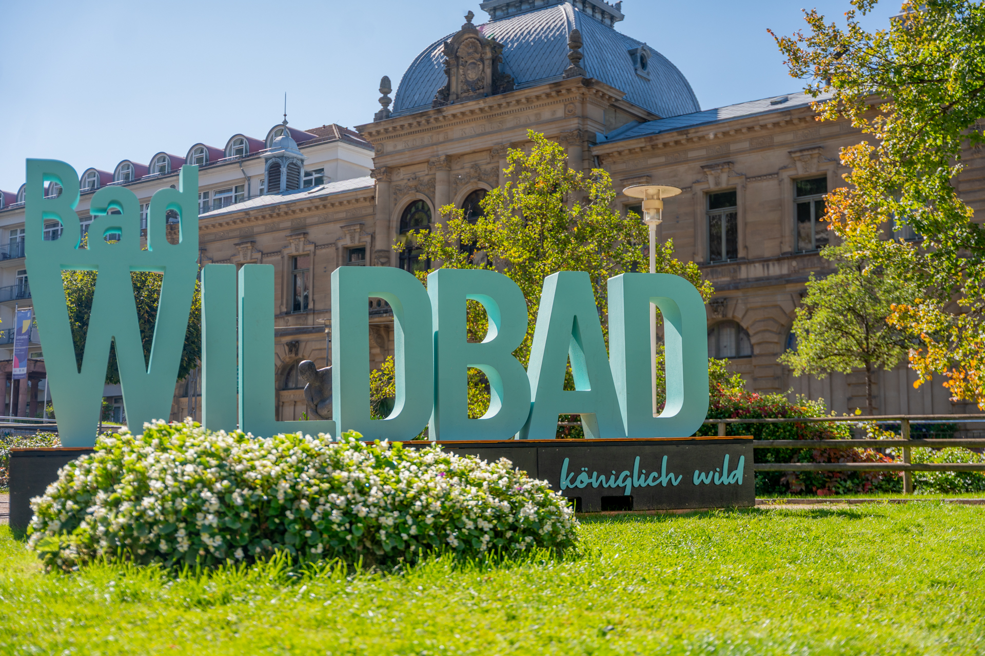Große grüne Buchstaben bilden den Schriftzug 'Bad WILDBAD' auf einer Wiese vor einem historischen Gebäude