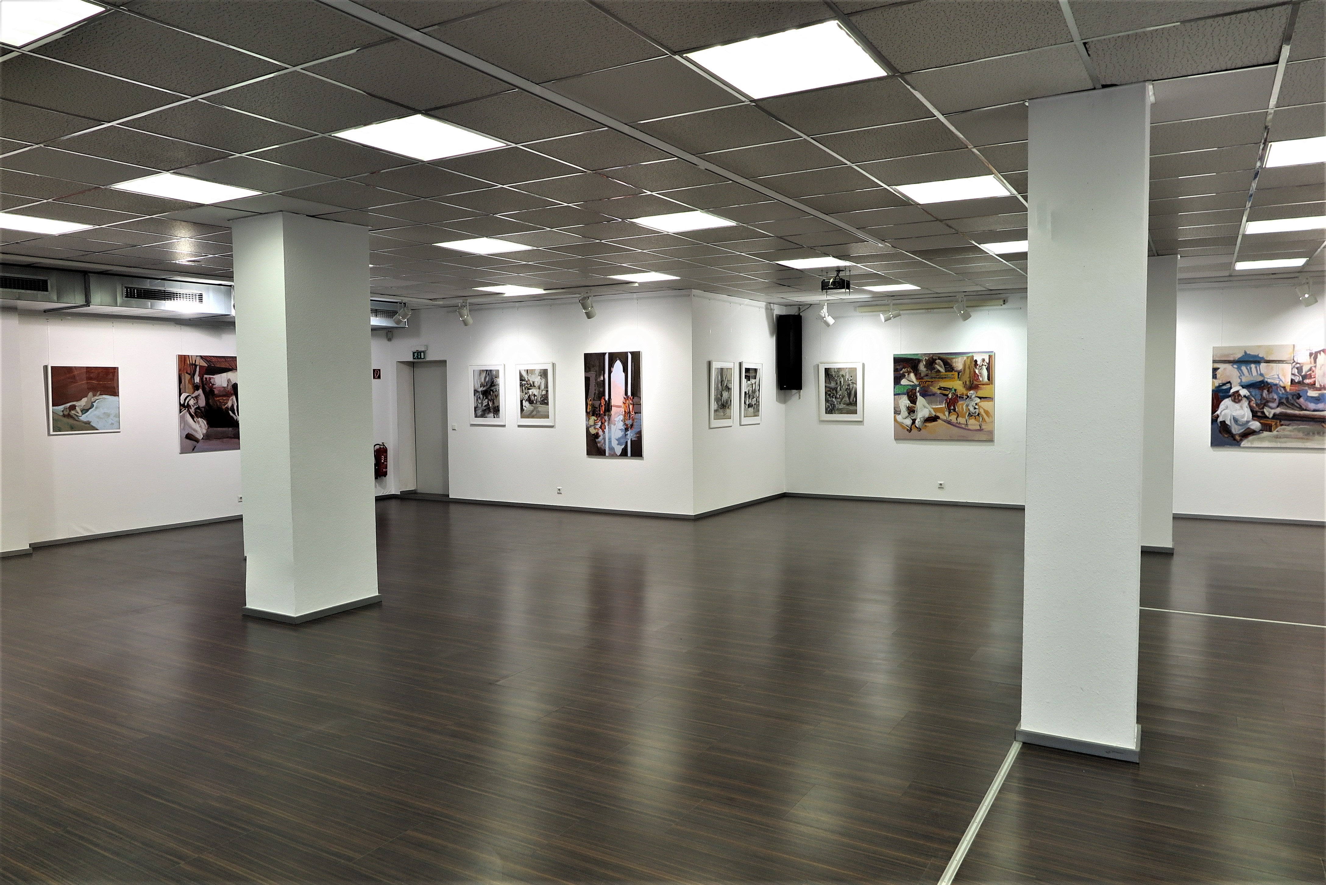 Innenansicht einer modernen Kunstgalerie mit weiß gestrichenen Wänden, dunklem Holzboden und Rasterdeckenbeleuchtung. An den Wänden hängen verschiedene Gemälde und Fotografien, die Szenen mit Menschen darstellen. Die Galerie ist geräumig, gut beleuchtet und wirkt ruhig und einladend.