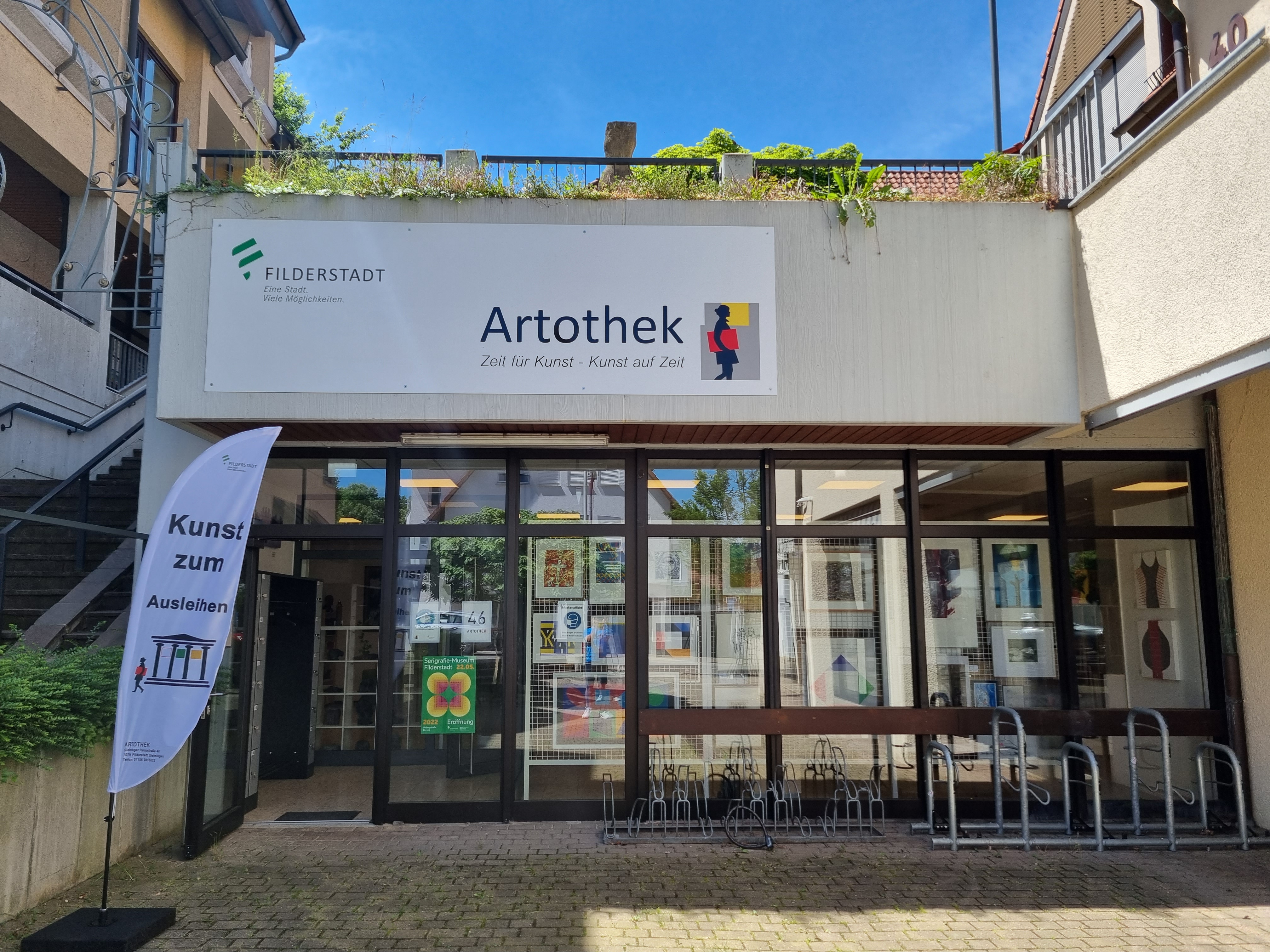 Eingangsbereich der Artothek Filderstadt bei sonnigem Wetter. Über dem Eingang hängt ein großes Schild mit dem Logo der Stadt Filderstadt und dem Schriftzug „Artothek – Zeit für Kunst – Kunst auf Zeit“. Links neben der Tür steht eine weiße Fahne mit der Aufschrift „Kunst zum Ausleihen“. Durch die Glasfront sind zahlreiche gerahmte Kunstwerke im Innenraum sichtbar. Vor dem Eingang befinden sich Fahrradständer.