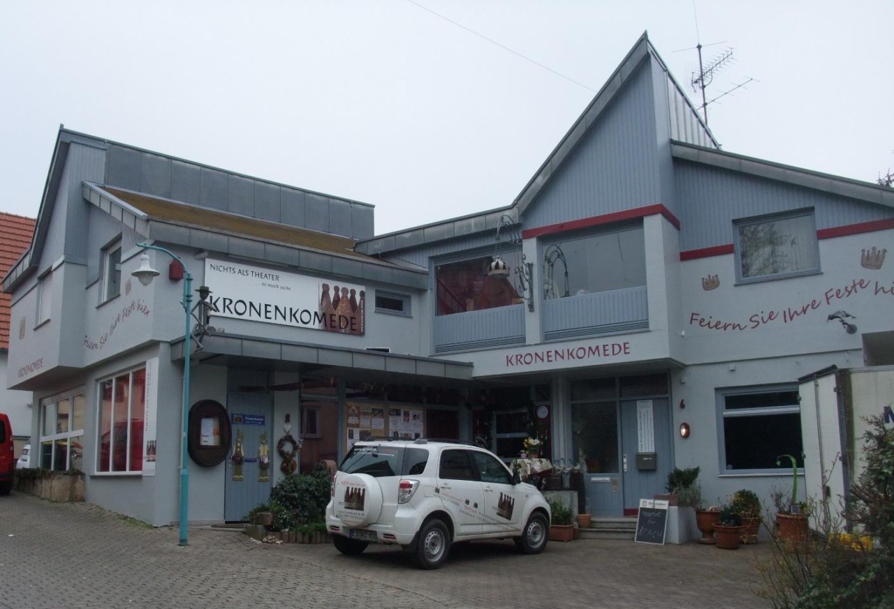 Außenansicht des Gebäudes „KronenKomede“ in Filderstadt, Deutschland. Es handelt sich um ein Theaterhaus mit der Aufschrift „NICHTS ALS THEATER“ und „Feiern Sie Ihre Feste hier“. Ein weißes Auto mit dem Logo des Theaters steht vor dem Eingang. Die Fassade ist hellgrau mit roten Akzenten, dekoriert mit Theatermotiven und Pflanzen.