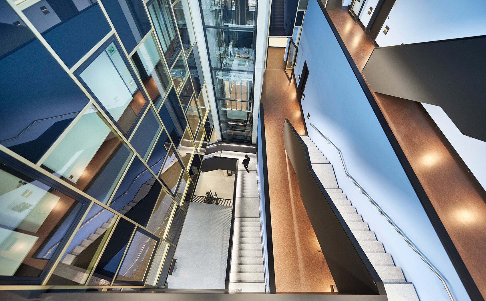 Blick von oben in ein modernes, mehrstöckiges Atrium mit Treppen, gläsernem Aufzugsschacht und Fluren.