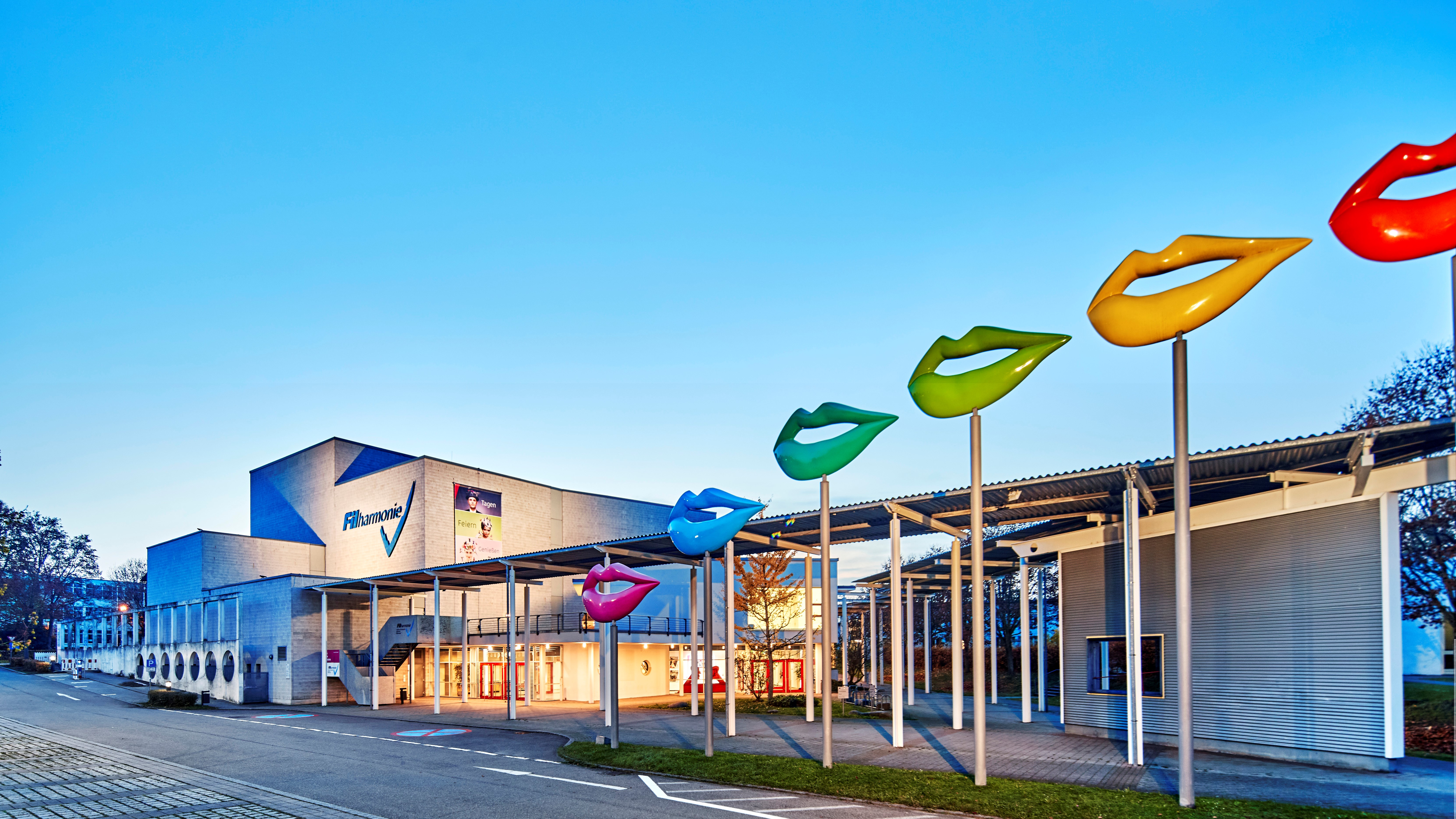 Aussenansicht der FILharmonie mit Blick auf den Eingang. Es dämmert, der Eingang ist hell erleuchtet. Vor den FILharmonie sind auf Metallpfosten in ansteigender Höhe modellierte Lippen in verschiedenen Farben angebracht.