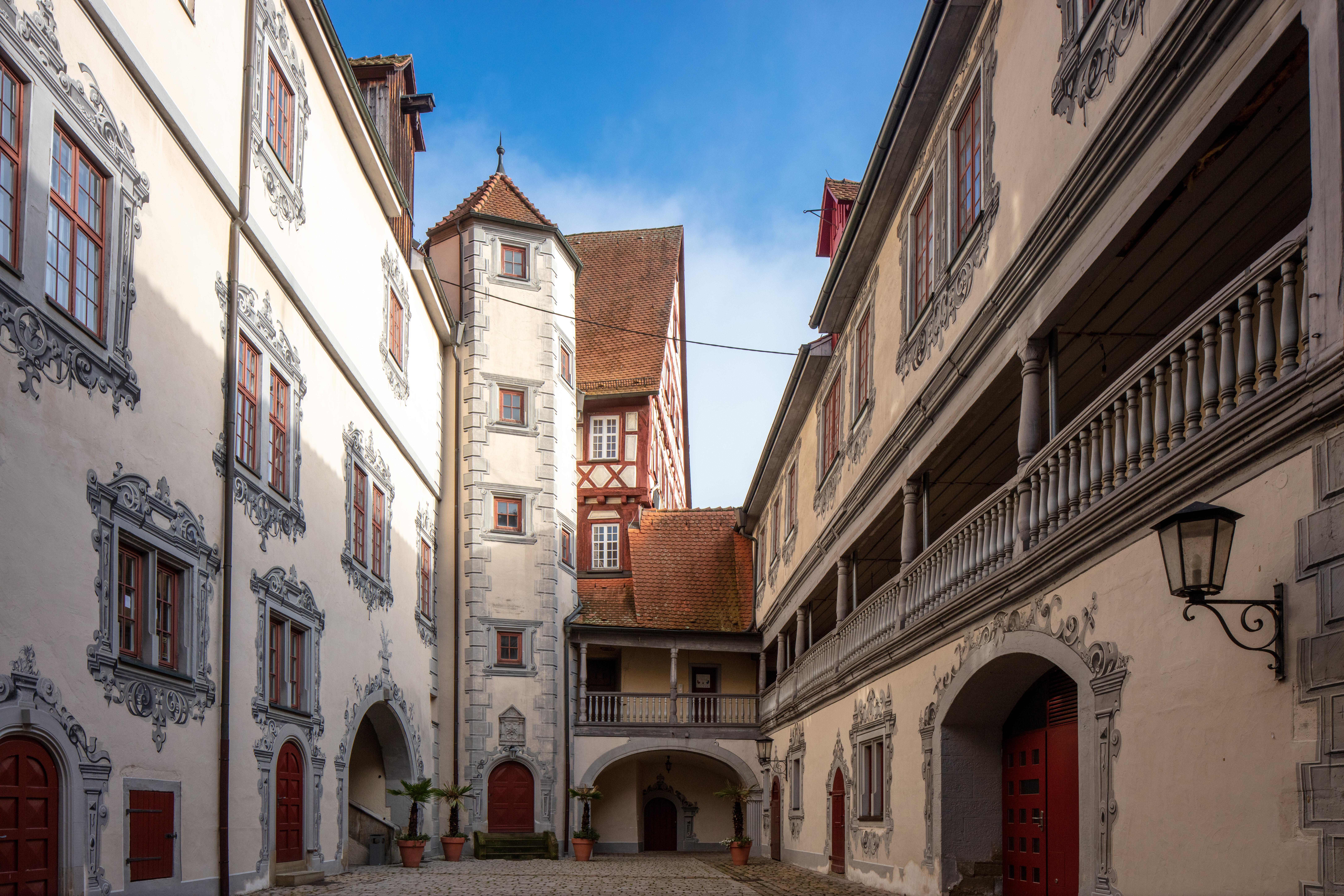Innenhof eines historischen Schlosses mit weiß verputzten Fassaden, roten Türen und Fenstern sowie einem blauen Himmel im Hintergrund
