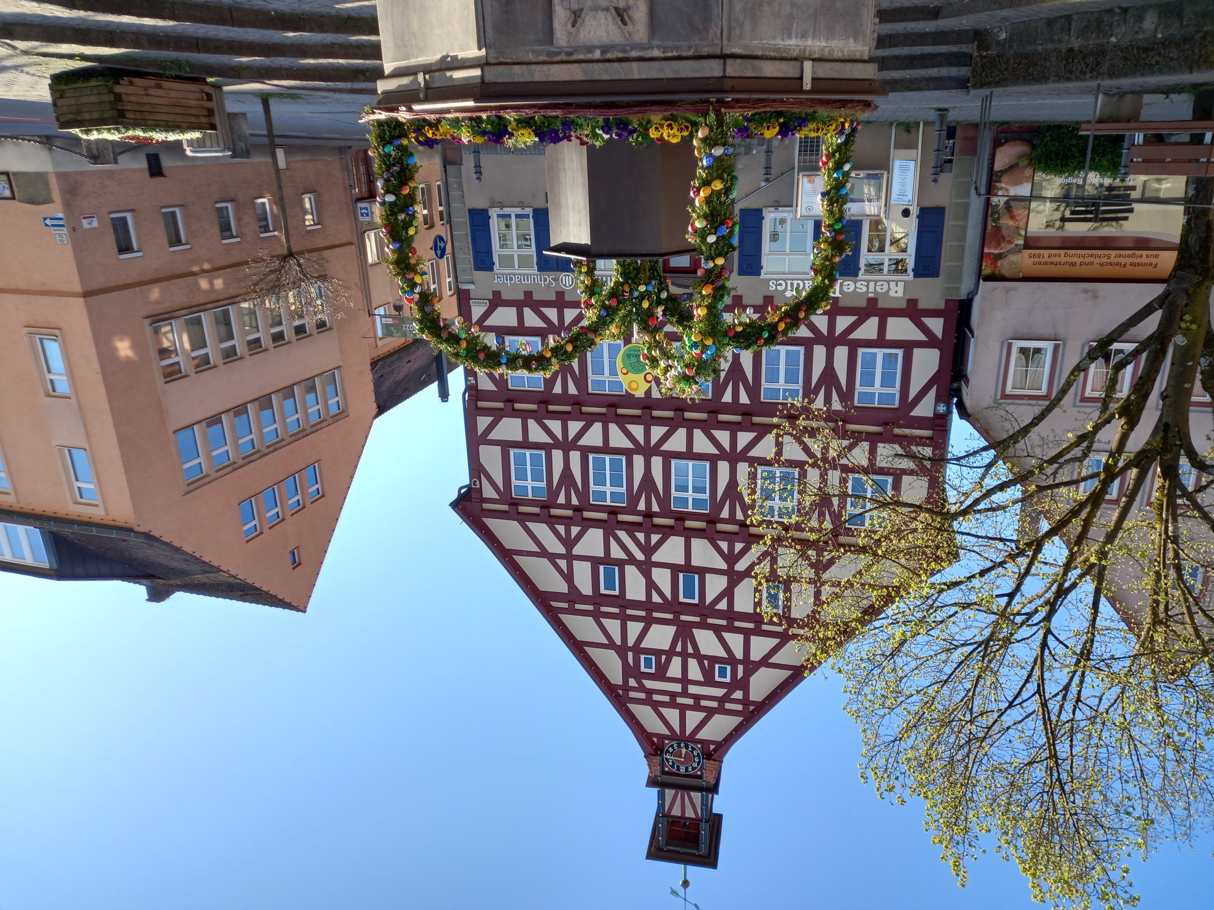 Fachwerkhaus mit Uhrturm hinter einem Brunnen, der mit Blumen und grünen Bögen geschmückt ist, an einem sonnigen Tag auf einem Marktplatz