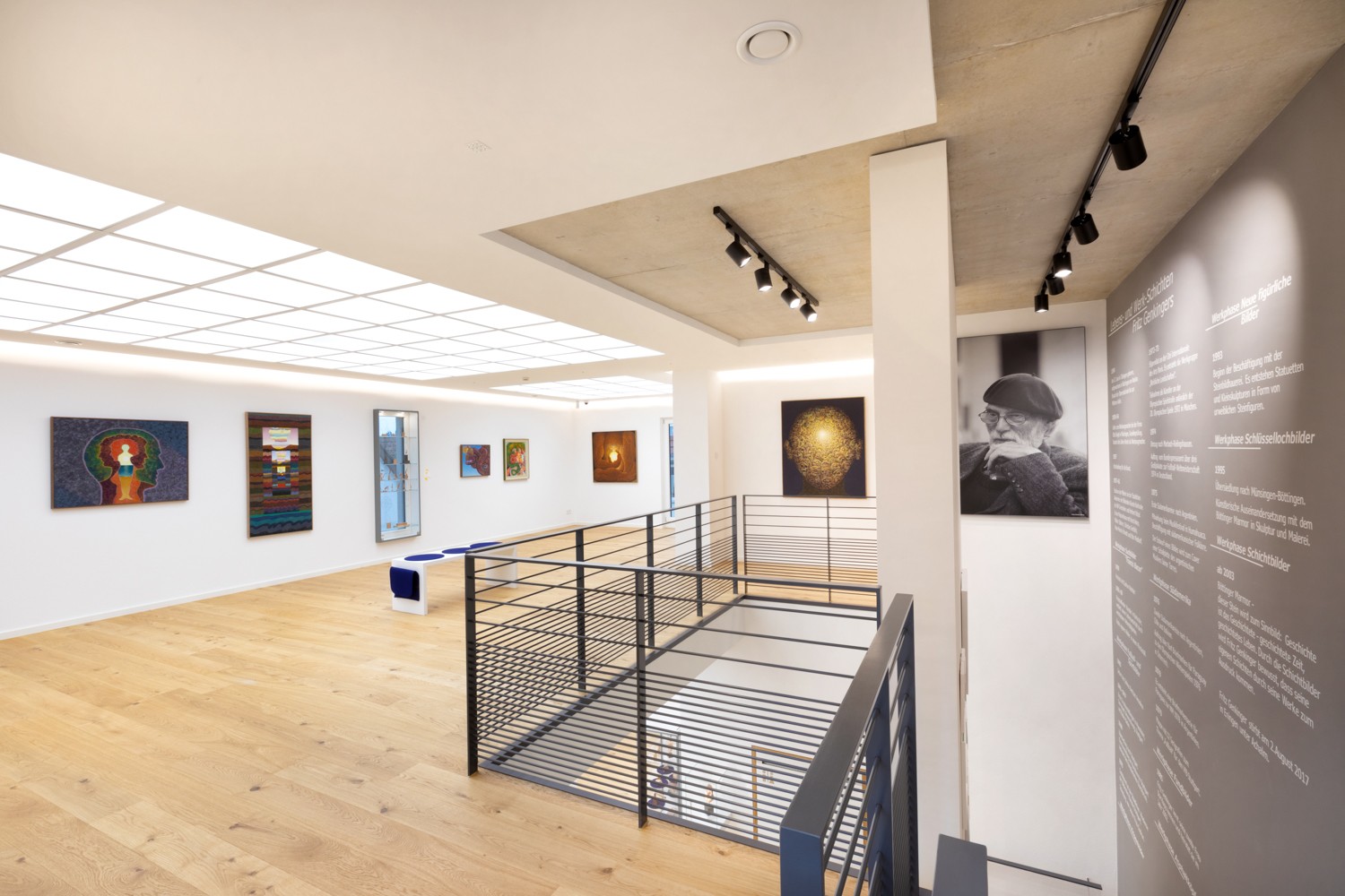 Innenraum einer Galerie im Fritz Genkinger Kunsthaus mit hellen Wänden, Holzfußboden und moderner Beleuchtung. An den Wänden hängen farbenfrohe Gemälde, im Vordergrund ein schwarzes Metallgeländer um das offene Treppenhaus. Rechts ein großes Porträtfoto von Fritz Genkinger sowie eine Wand mit biografischem Text über den Künstler.