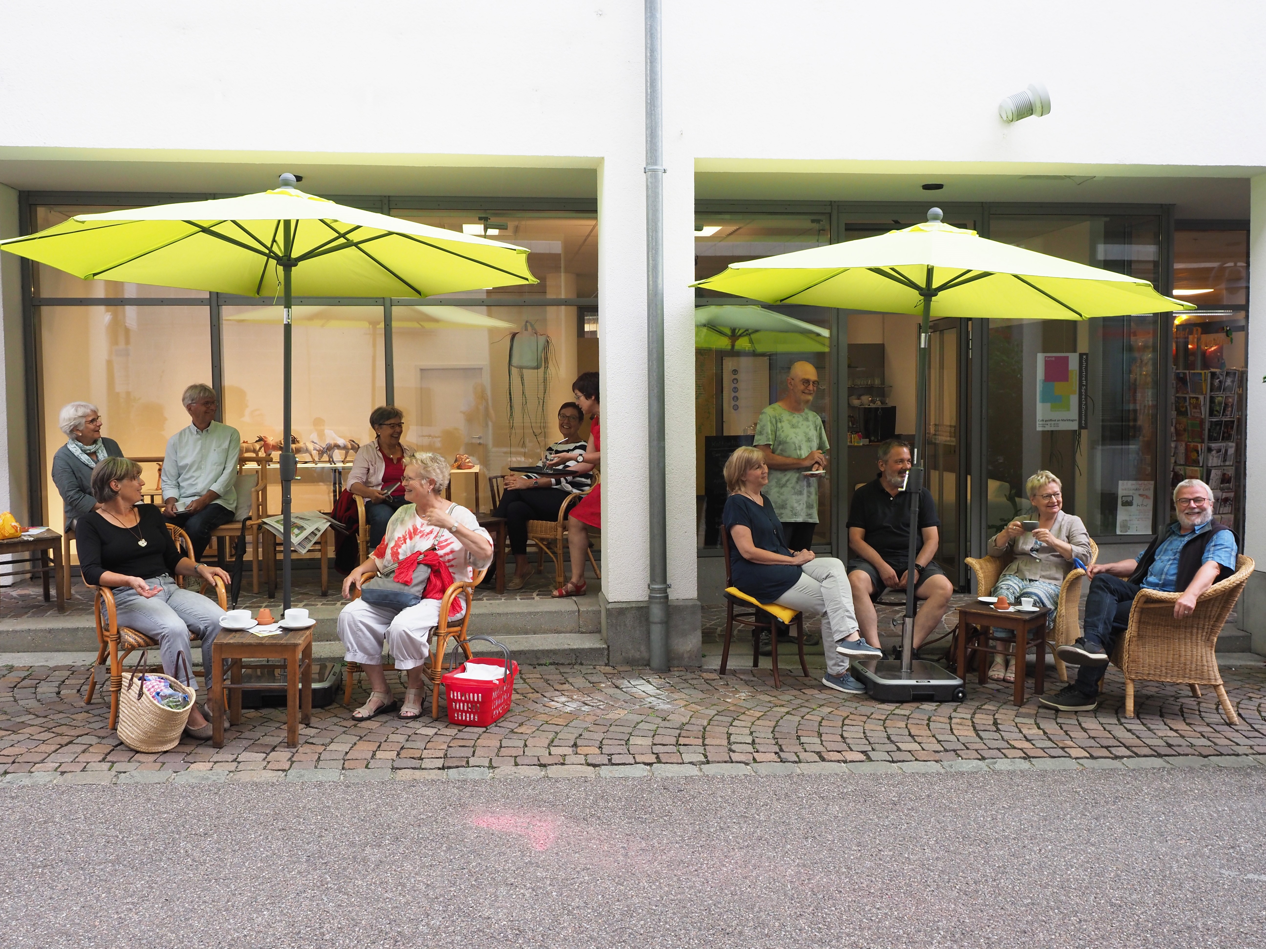Menschen sitzen unter Sonnenschirmen vor den Café SprechZimmer