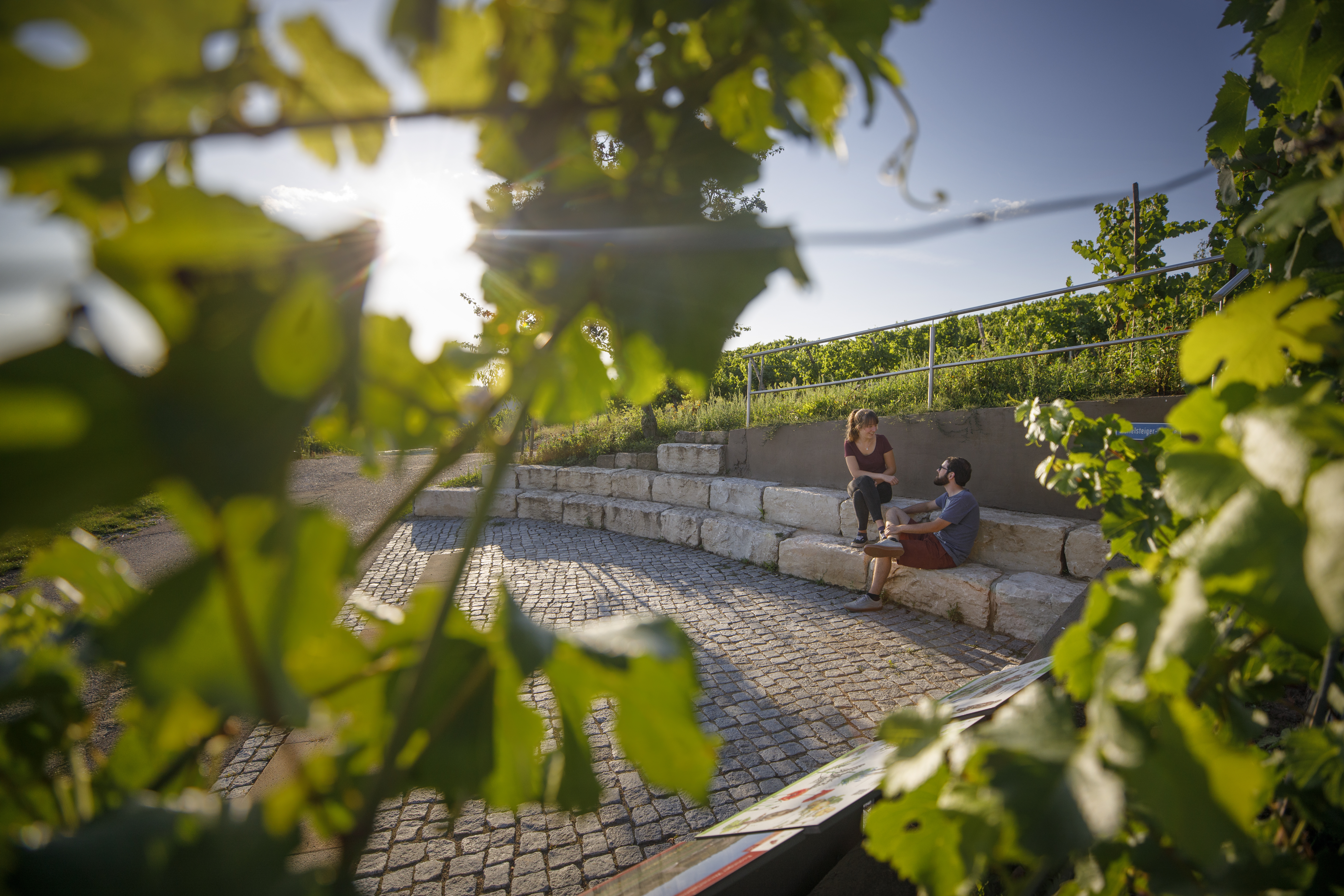 Zwei Personen, möglicherweise ein Paar sitzen auf halbrund angeordneten Steinen. Es wurde durch eine Weinrebe hindurch fotografiert, so dass im Vordergrund am Bildrand noch verschwommen Blätter erkennbar sind