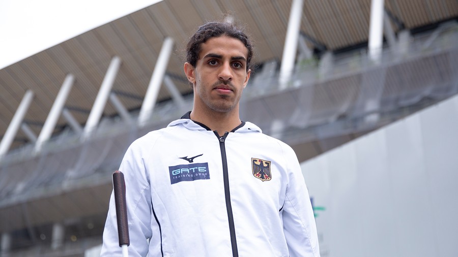 Junger Mann mit dunklen Haaren in weißer Sportjacke mit deutschen Wappen und Sponsorenlogos vor moderner Architektur