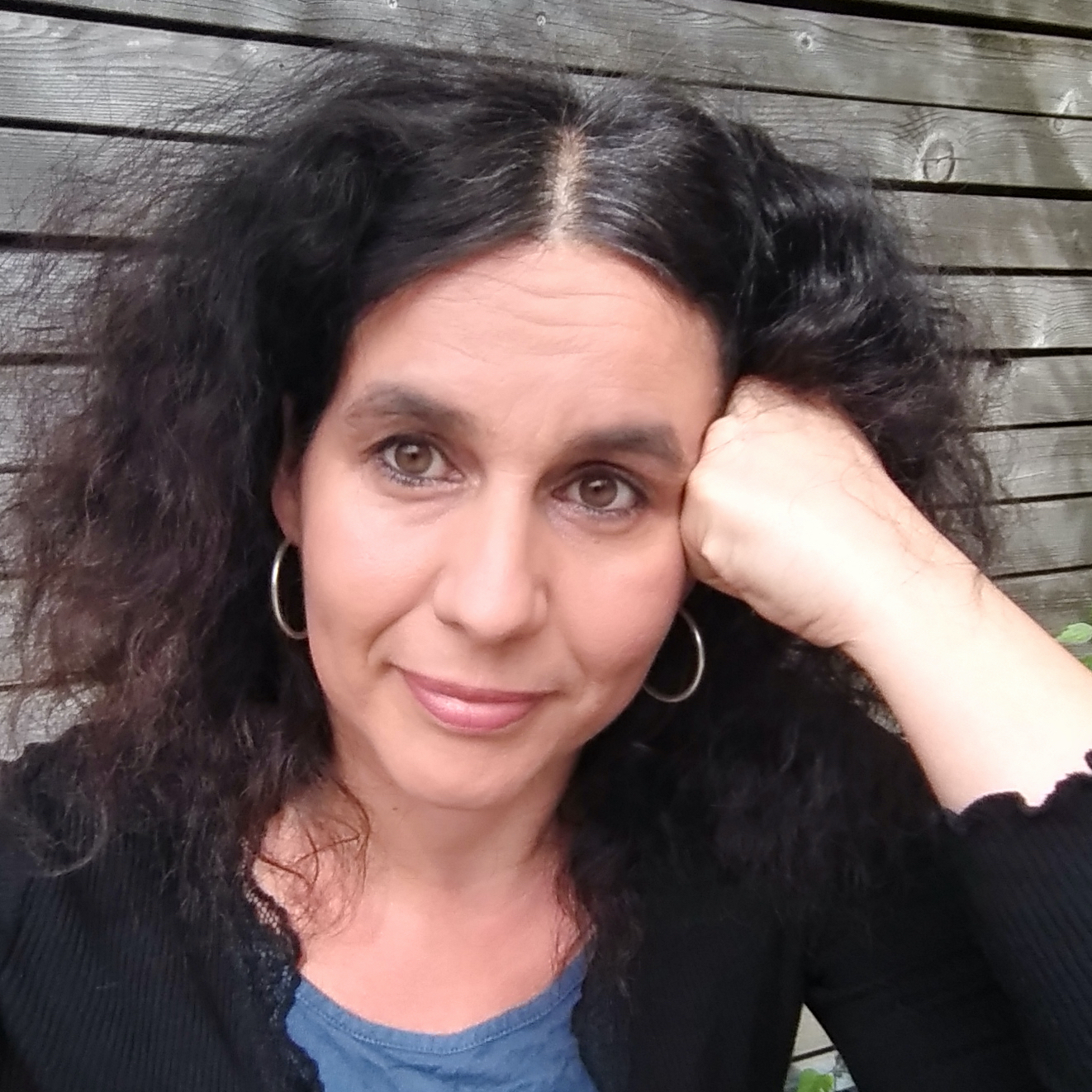 Portraitbild von Ramona Ambs. Ramona Ambs hat langes, dunkles, lockiges Haar und braune Augen. Auf dem Bild trägt sie eine schwarze Bluse oder Jäckchen über einem blauen Shirt und große silberne Creolen-Ohrringe. Sie blickt mit einem angedeuteten Lächeln in die Kamera und stützt den Kopf auf eine Hand.